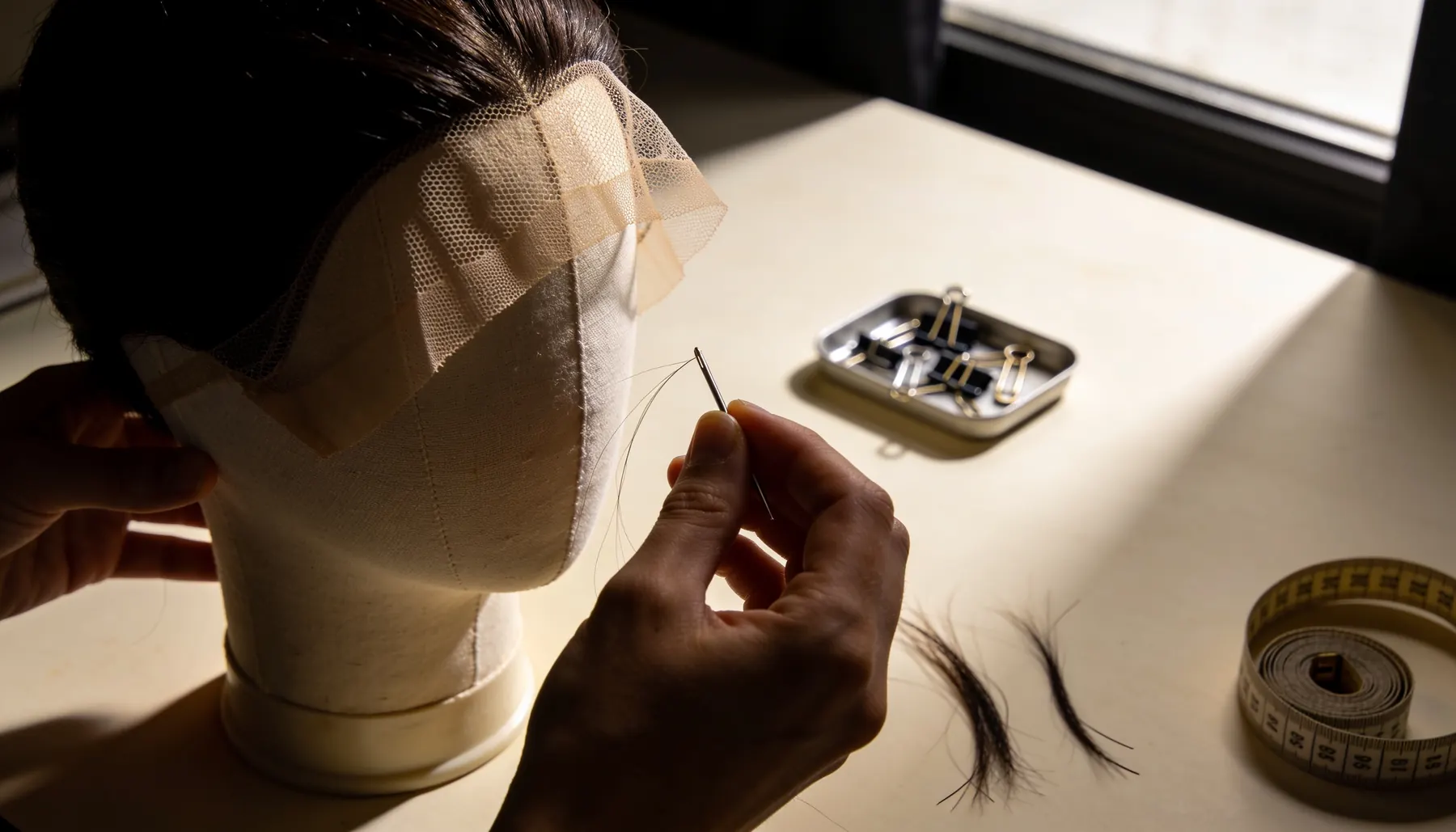 Close view of a hand ventilating hair into a fine lace base on a canvas block, with clips and a measuring tape on the bench.